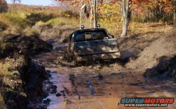 Truck struggling through a muddy off-road trail with trees and fall foliage in the background