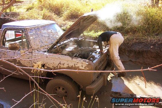 Mud-covered pickup truck with hood open, steam rising from engine, stuck in mud