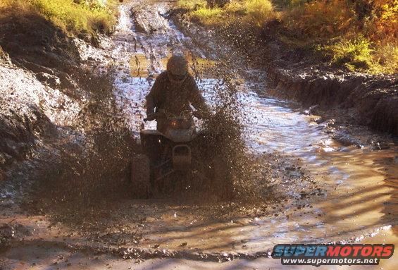 ATV driving through a muddy trail, kicking up a lot of water and dirt
