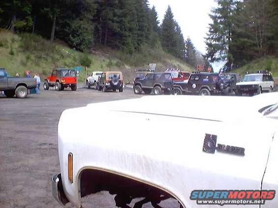 Several Jeeps and a pickup truck parked on a gravel lot with trees in the background. A white truck is in the foreground