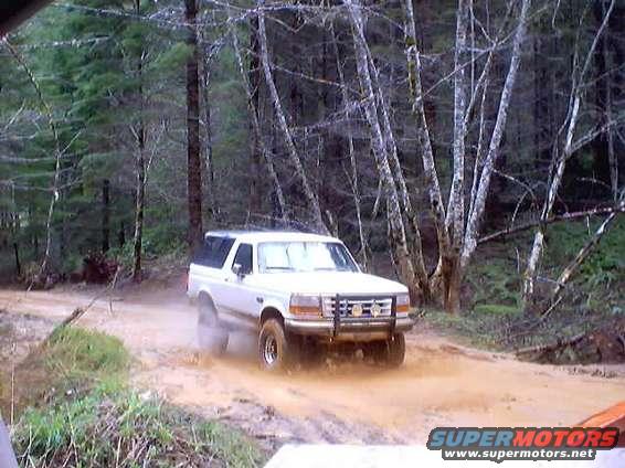White Ford Bronco driving through mud on a dirt road in a wooded area