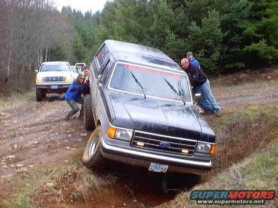 Ford truck stuck in a ditch with people pushing it. Another truck is behind it