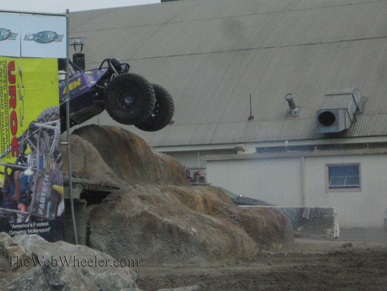 Purple off-road buggy mid-air over a dirt ramp. Building in background