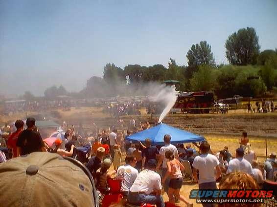 People gathered around a blue tent while a water spray shoots up from it. A red truck is in the background