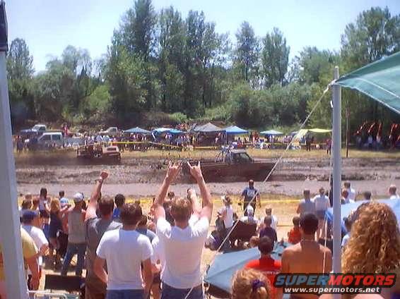 Muddy track with several vehicles, spectators, and tents under a bright sky