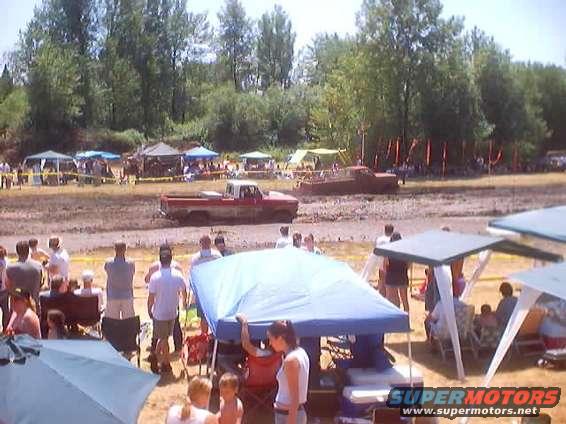 Red pickup truck in mud, side view. Muddy ground, trees, tents, and people in background