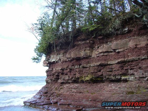 Rock cliff face with layered strata, trees on top, and water below