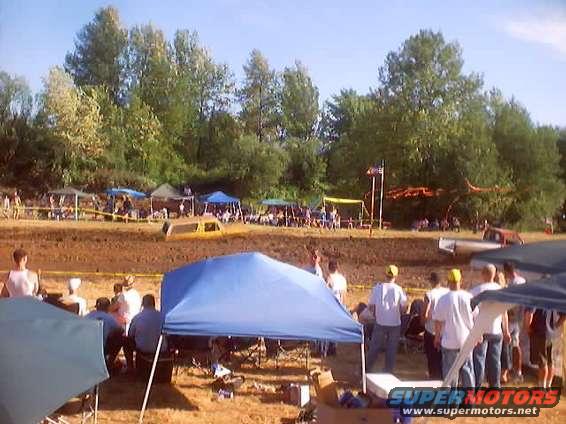 Yellow pickup truck stuck in mud, side view, with trees and tents in background