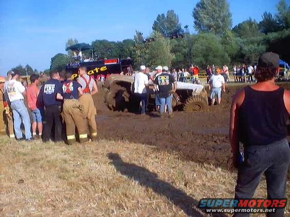 People watching a mud bogging event with a truck stuck in the mud. A large black and red vehicle is visible in the background