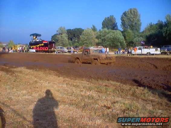 Mud-covered 1976 Ford F-100 driving through a mud pit, with a "Pirate Radio" stage in the background