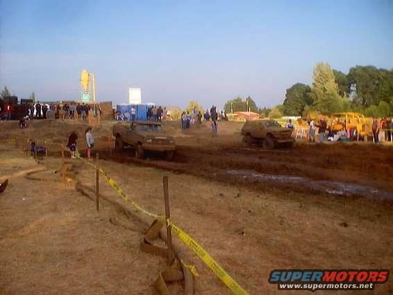 Two trucks are in a mud pit with spectators watching. One truck is in the foreground, the other is further back