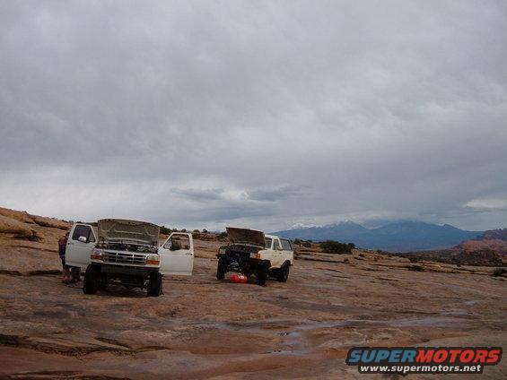 Two white SUVs and a dark SUV parked on rocky terrain, hoods up, with mountains in the background
