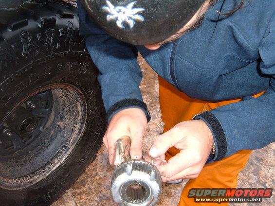 Person in blue jacket and orange pants works on a metal vehicle part near a large tire
