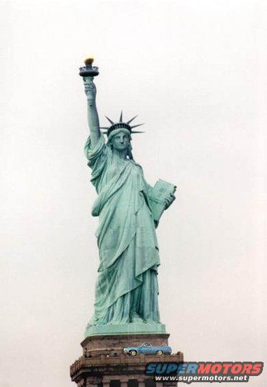 Statue of Liberty with a small blue car on its pedestal, Supermotors.net logo visible