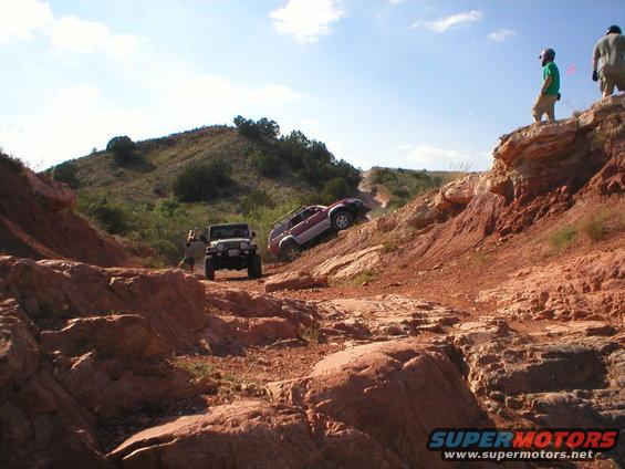 Jeep and red SUV climbing a steep, rocky off-road trail with two people watching from a ledge