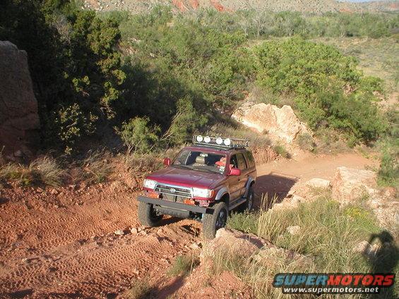 1996 Toyota 4Runner driving on a dusty, rocky trail with scrub brush and red rock formations in the background
