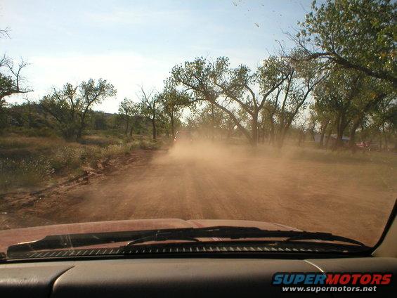 Driving down a dirt road kicking up dust, trees on the side, other vehicles visible ahead