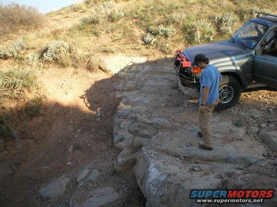 Man in blue shirt and khaki pants stands by a dark gray SUV with a brush guard, parked on rocks near a dirt drop-off. SUV appears to be a 1998 Toyota Land Cruiser