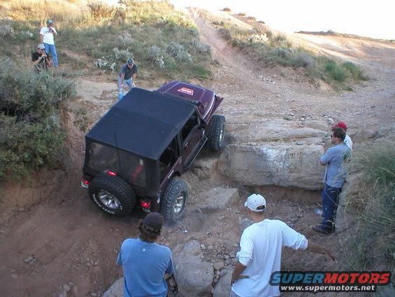 Dark purple Jeep Wrangler with a black soft top navigating a rocky off-road trail, viewed from the rear-left