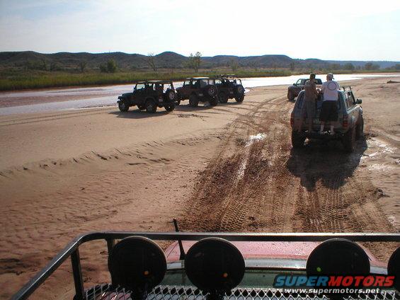 Several off-road vehicles, including Jeeps and a pickup truck, driving on a sandy, muddy riverbed with hills in the background