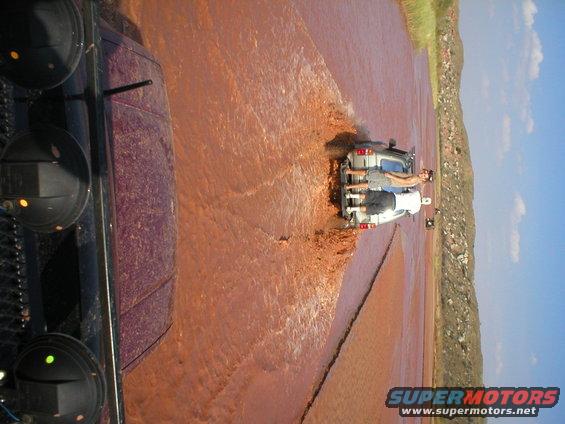 Silver SUV driving through muddy water, rear view. Two people on the back