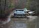 1978 Ford Bronco - A blue Ford Bronco navigates a flooded path, with a damaged vehicle partially visible behind it.