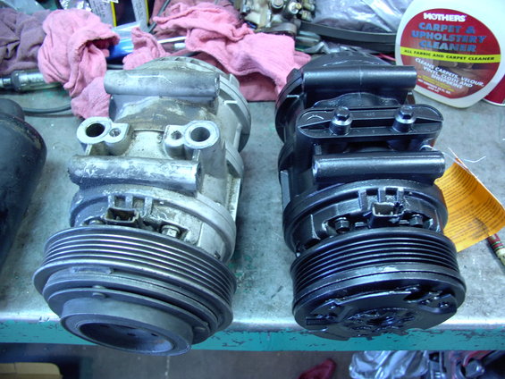 Two AC compressors, one old and corroded, the other new and black, sit on a workbench next to a bottle of Mothers carpet cleaner