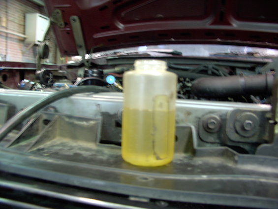 Plastic fluid container with yellow liquid sits in the engine bay of a dark red vehicle with the hood open