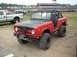 1978 Ford Bronco - A modified red SUV with large off-road tires and a winch is parked on a dirt lot, with a stadium in the background.