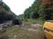 1978 Ford Bronco - A dark pickup truck is stuck in a muddy path surrounded by trees, while a yellow vehicle is partially visible in the foreground.