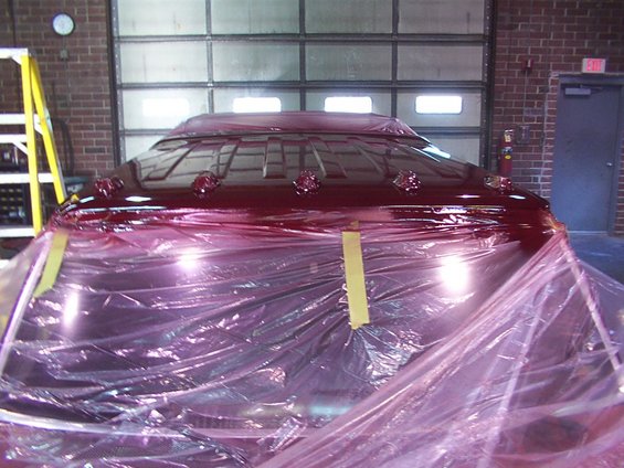 Dark red vehicle roof with raised sections, covered in pink plastic sheeting. Garage interior with overhead door and ladder visible