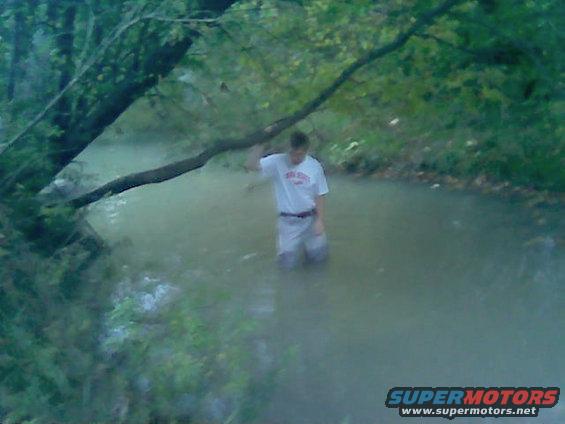 Person wading in a stream with a tree branch overhead. Watermark reads SUPERMOTORS