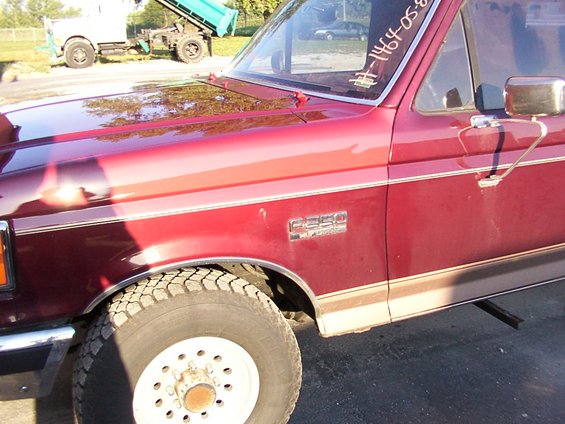 Red Ford F-250 pickup truck, side view showing fender badge and two-tone paint. Truck has a white wheel and knobby tire. A dump truck is in the background