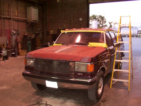 Maroon Ford F-Series truck with red hood and yellow tape on windshield, parked in garage with ladder