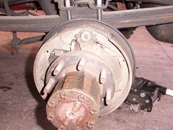 Close-up of a rusty rear drum brake assembly with lug nuts and part of a leaf spring visible