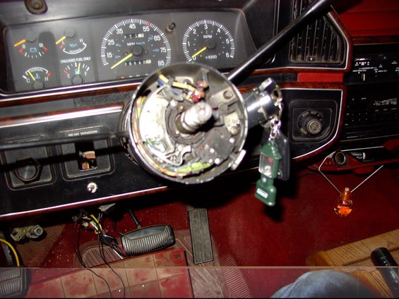 Interior shot of a vehicle's steering column with exposed wiring and keys. Dashboard gauges and radio visible