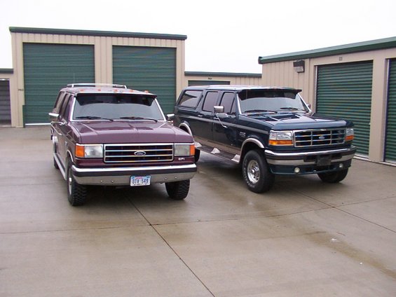 Two Ford Bronco Centurion Conversions parked in front of storage units. The one on the left is maroon, and the one on the right is dark blue