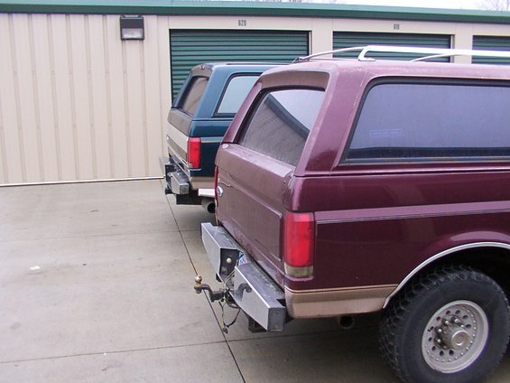 Two Ford Bronco SUVs parked in front of storage units. The one in front is maroon, and the one behind it is dark green