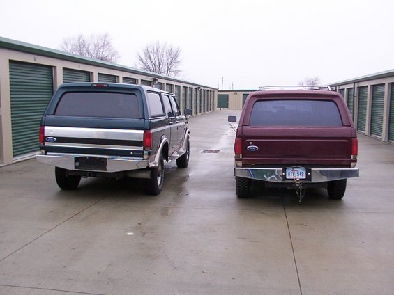 Two Ford Broncos with camper shells parked in front of storage units. The one on the left is dark green and silver, the one on the right is maroon