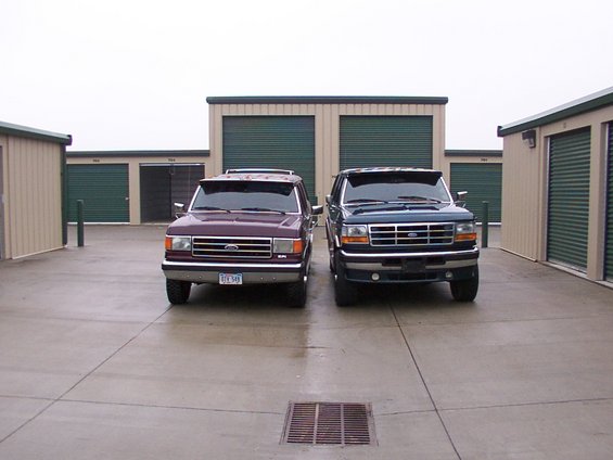 Two Ford trucks, one maroon and one blue, parked front-facing in front of storage units. The blue truck appears to be a 1992 model