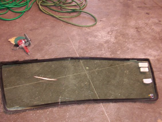 Windshield with cracks and a thin white object on it, lying on a concrete floor next to a grinder and green hoses