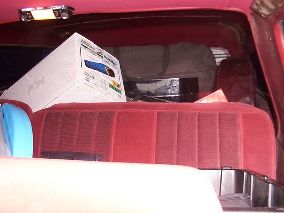 Interior of a red velour-clad vehicle, showing a sleeping bag box and duffel bag on the rear seat