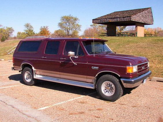 Maroon Ford F-Series SUV conversion with tan trim, parked in gravel lot