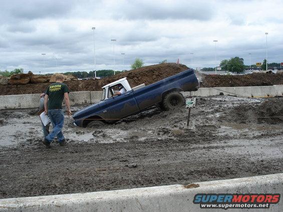 Blue pickup truck stuck in mud pit with two people standing nearby