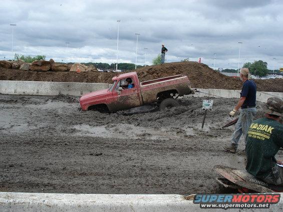 Red pickup truck stuck in mud, driver visible, tow rope attached