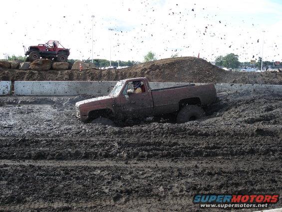 Red pickup truck stuck in mud pit, driver visible, another off-road vehicle on embankment