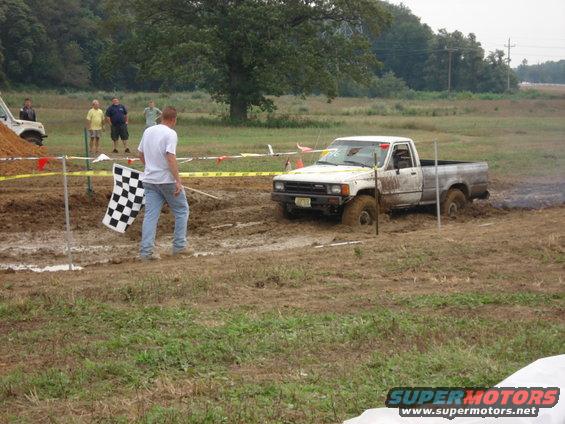 White pickup truck stuck in mud, with a person holding a checkered flag nearby. Another white SUV is visible in the background