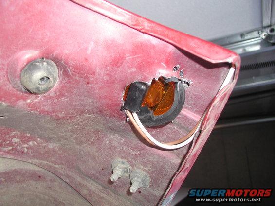Close-up of a damaged amber marker light assembly on a red vehicle's body panel. Wires are visible, and the plastic lens is broken