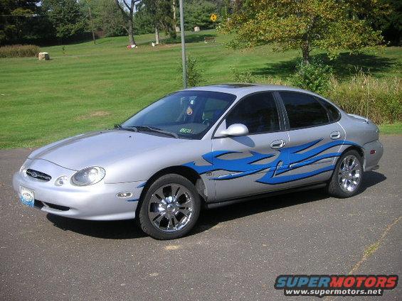 Silver 1999 Ford Taurus sedan with blue flame decals and chrome wheels, parked on asphalt
