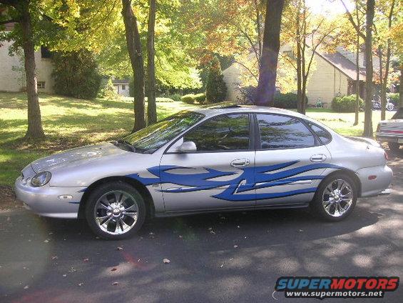 Silver 1999 Ford Taurus sedan with blue flame decals and chrome wheels, parked on asphalt